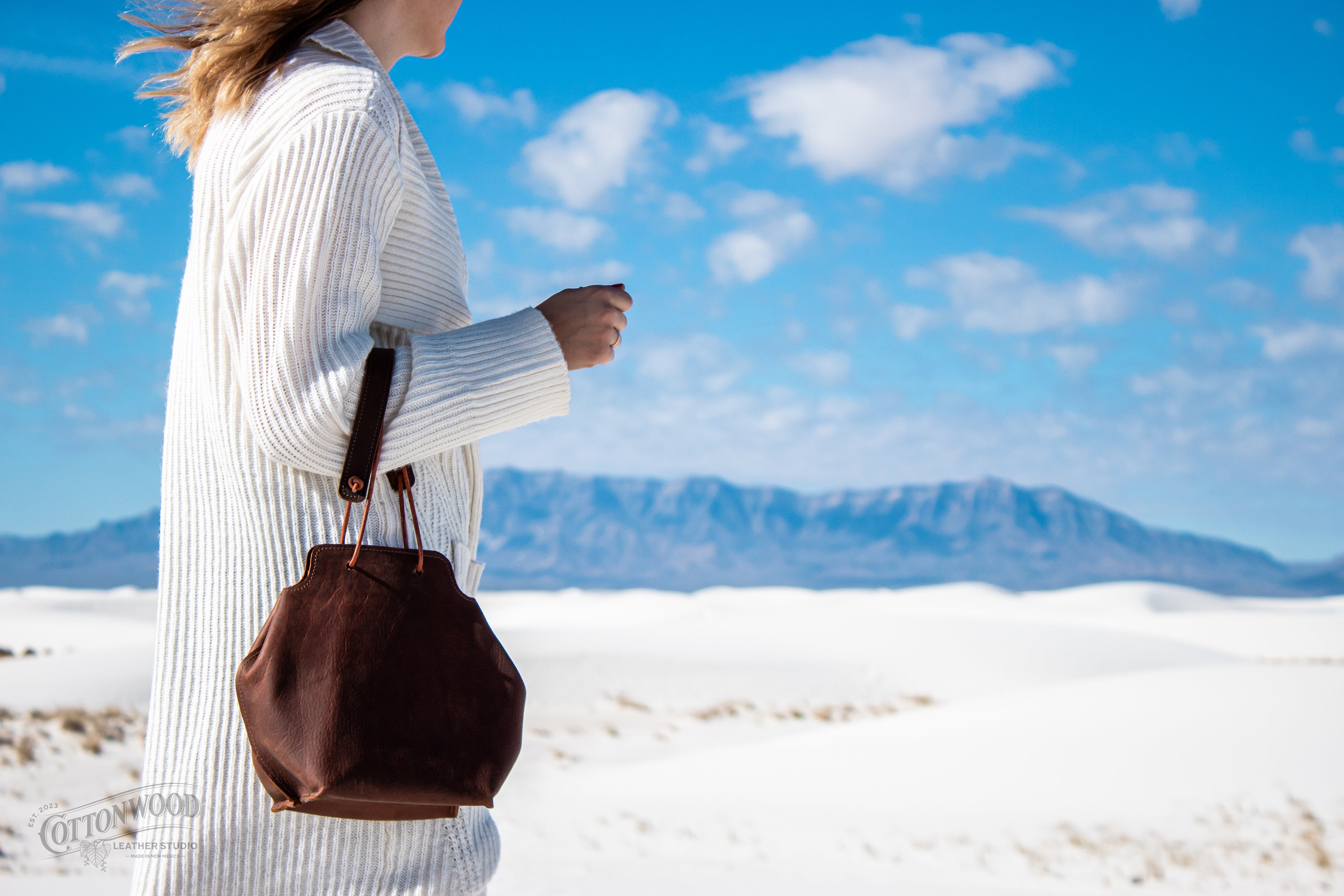 Woman walked on the sand with mountains in the background carrying a brown leather purse