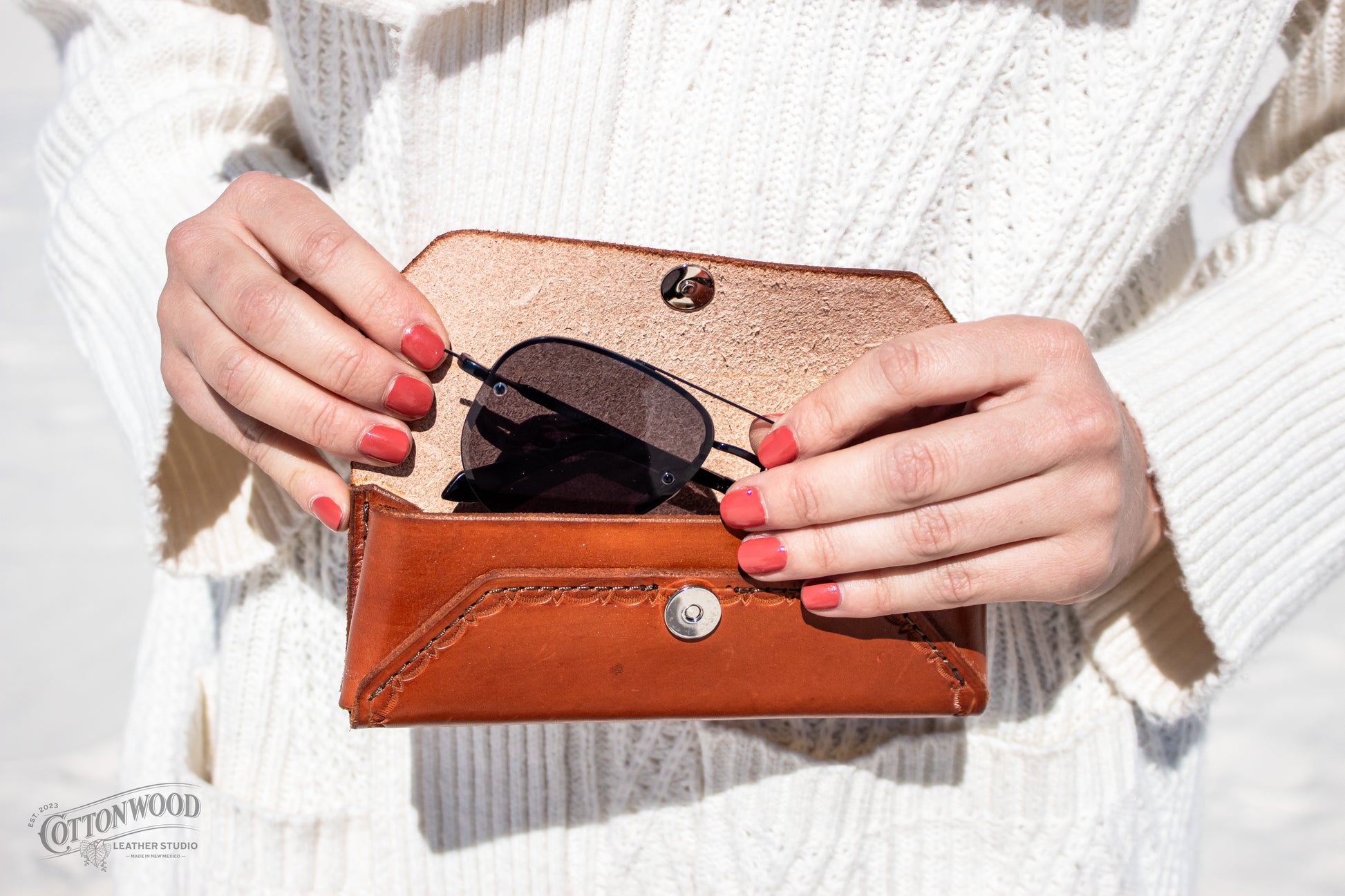 Hands holding open a golden brown leather glasses case with sunglasses peaking out